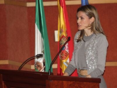 La Reina Letizia de España pronuncia un discurso en una conferencia, con la bandera española y otra nacional al fondo.