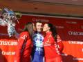 Un ciclista en un traje azul y blanco celebra con dos personas vestidas de rojo, uno de ellas sosteniendo un trofeo. El fondo es una pared con publicidad de Cruzcampo.