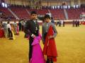 Dos jóvenes vestidos con trajes de torero posan en un escenario deportivo, rodeados por otros participantes y espectadores.