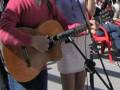 Un hombre con una camisa roja y una mujer con un vestido blanco están cantando en un escenario. El hombre toca la guitarra mientras la mujer canta en el micrófono. La escena parece ser una actuación musical al aire libre, posiblemente en un evento o fiesta.
