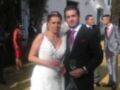 A una pareja casada posando en un evento formal, con la novia en un vestido blanco y el novio en traje negro. La escena se desarrolla frente a un edificio blanco con detalles arquitectónicos, y otros invitados están presentes en el fondo.