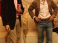Dos hombres jóvenes posan en un entorno elegante, uno con chaqueta negra y pantalones beige, el otro con chaqueta marrón y jeans. Ambos parecen estar en una presentación o evento formal, con detalles decorativos en el fondo.