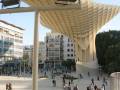 Imagen del Metropol Parasol en Sevilla, España. Arquitectura moderna con estructura de madera y acero. Paseo público con gente caminando y sentada en el parque. Edificios blancos al fondo bajo un cielo azul claro.