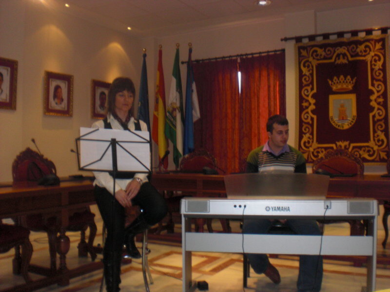 En una sala de conferencias, un hombre toca el piano mientras otra persona se sienta en el fondo. La sala está decorada con banderas y un escudo heráldico.