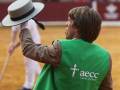 Un hombre con una chaqueta verde de AECC, un sombrero y un palo en la mano, posa en una pista de arena.