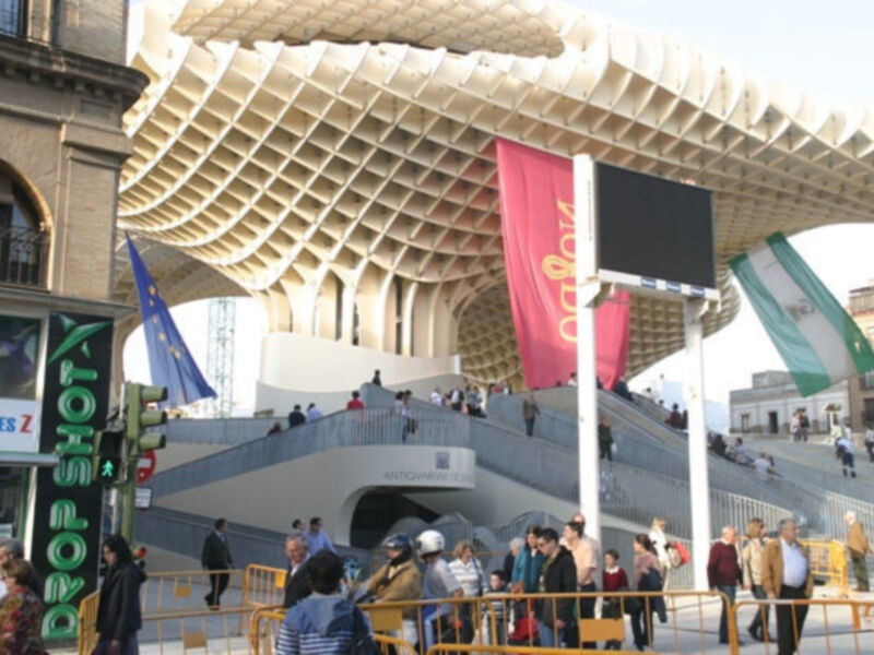 Una estructura moderna con un techo de malla, posiblemente el Metropol Parasol en Sevilla. Debajo hay una escalera con gente caminando y varios carteles publicitarios, incluyendo uno que dice "Deportes Z". La escena parece ser en un día soleado con cielo azul.