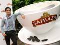 Un hombre sonríe mientras posa junto a una gran taza de café gigante con el logotipo Saimaza. En la base de la taza, hay algunos granos de café.