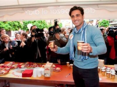 Persona celebrando con vasos de café en un evento público, rodeada de espectadores y cámaras de fotos.