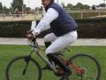Un hombre monta una bicicleta en un campo de césped, con otros asistentes y estructuras al fondo.