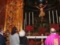 En una iglesia, una imagen de Cristo crucificado se encuentra en un altar dorado. A su lado, varias personas observan la escena religiosa. En el fondo, hay flores y velas encendidas en una mesa blanca.
