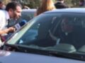 Un hombre con una camisa blanca está hablando a un hombre en el asiento del conductor de un coche azul. El hombre en el asiento del conductor lleva gafas y una camisa negra con un cuello de sacerdote. Al fondo, hay personas observando la escena.