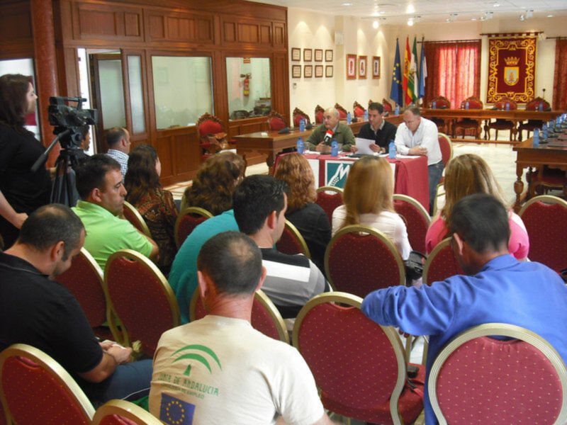 Una sala de conferencias con una mesa central donde se reúnen varios hombres en trajes, junto a un hombre en camisa blanca. La sala está decorada con banderas y estandartes, y hay personas sentadas en sillas rojas escuchando a los hombres en la mesa.
