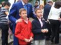 Un hombre en un traje azul y corbata se encuentra con dos niños, uno vestido de rojo y otro con chaqueta azul. Ambos sostienen pequeños trofeos, posiblemente en una celebración o evento. Al fondo, se observan otros participantes y un edificio con balcones.