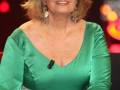 Una mujer con cabello rubio corto y ondulado, lleva un vestido verde de satén con detalles en la cintura. La imagen parece ser de una transmisión televisiva, con luces y efectos en el fondo.