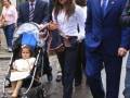 Una familia camina por una calle, con un hombre en traje azul y una mujer con vestido blanco. La niña está sentada en un carrito de bebé, y otros adultos están a su lado.