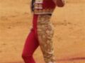 Un torero en traje tradicional de color rojo y dorado, con guantes negros y un sombrero, sostiene una vara de mando en la mano izquierda. El fondo es el suelo rojizo característico del ruedo de toros.