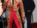 Un torero en traje tradicional de color rojo y dorado, con una corbata negra, posa para la cámara mientras un hombre en uniforme lo observa.