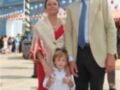 Una familia feliz en un parque de atracciones, con una madre vestida en traje rojo flamenco, padre con traje beige y niño con camisa blanca y pantalones cortos. En el fondo, una gran rueda de la fortuna y banderas ondeando en un día soleado.