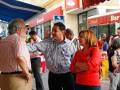 Fotografía de una calle con gente caminando y conversando. En el fondo, se puede ver un edificio con una tienda de barbacoa y una bandera. La gente está vestida casualmente, con camisas de rayas y pantalones.