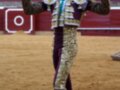 Un torero en un entorno de corrida, con traje tradicional dorado y morado, posando en el centro del ruedo. La arena está llena de espectadores detrás de una cerca roja y blanca con un anillo blanco en el centro.