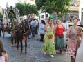 Un grupo de personas camina por una calle adoquinada, seguidas por un carro tirado por dos caballos. Las mujeres llevan vestidos tradicionales y sombreros verdes, mientras que los hombres están vestidos con camisas de manga larga. La escena parece ser parte de una celebración o fiesta cultural local.