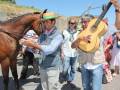 Un hombre con sombrero y chaleco azul está montando un caballo marrón. Otros individuos, incluyendo una mujer con guitarra acústica y otra con vestido blanco, observan la escena. La imagen parece ser de un evento cultural o tradicional en un entorno rural con una carretera y vegetación al fondo.