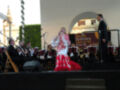 Una cantante en un vestido blanco y rojo, frente a una orquesta de músicos. La escena parece ser un concierto al aire libre con una bandera que dice "Banda Sinfónica Municipal Sevilla" en el fondo.