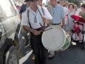 Un hombre con sombrero y gafas de sol toca un tambor en una calle. Detrás, otros personas caminan y observan la escena. El ambiente parece ser de una celebración o evento público.