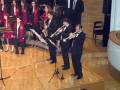 Un grupo de músicos en trajes formales toca instrumentos como saxofón, trompeta y clarinete en un escenario con una orquesta de fondo. La imagen captura la energía y el entusiasmo del concierto, destacando la concentración de los músicos y la atmósfera musical.