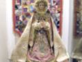 Imagen de una Virgen con vestimentas doradas y rosadas, posiblemente en un altar. Al fondo hay una tapicería de colores vivos, y a los lados se ven partes de otras imágenes religiosas.