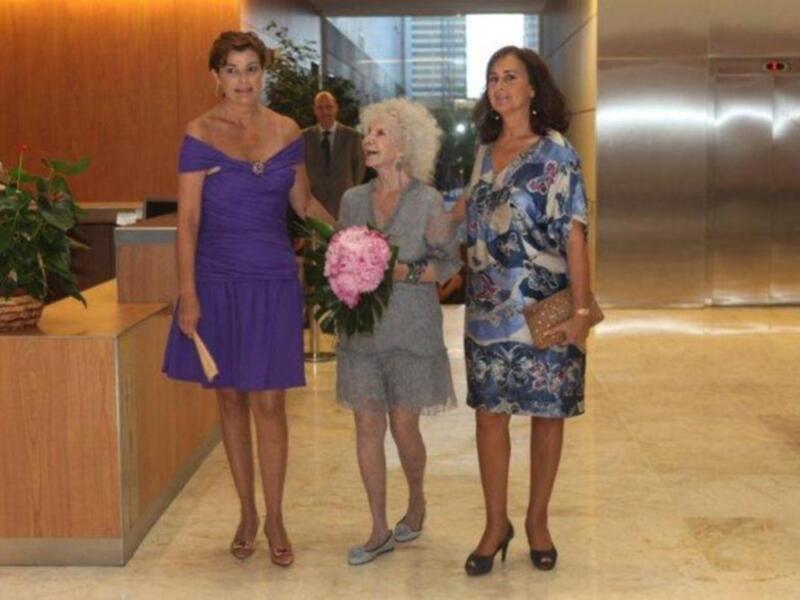 Tres mujeres posan en un vestíbulo de oficinas, dos con vestidos azules y una con un vestido gris claro. La imagen incluye elementos de lujo como un ascensor y una recepción con plantas.