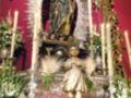 Un altar religioso con una estatua de la Virgen María en el centro, rodeada por flores y velas encendidas. La imagen es de una iglesia con detalles dorados en el arco y fondo rojo.