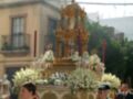 Fotografía de una procesión religiosa en un entorno urbano, con una imagen central de una estatua o imagen sagrada rodeada por flores y decoración dorada. A la izquierda, un hombre con uniforme rojo y a la derecha, otro con traje militar. En el fondo, edificios típicos de la ciudad.