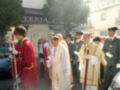 Imagen de una procesión religiosa con sacerdotes vestidos en rojo y blanco, acompañados por soldados uniformados. La escena se desarrolla frente a una tienda llamada "Ferretería".