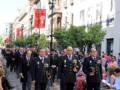 Desfile militar con uniformes ceremoniales, banderas y faroles en la calle.