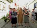 Un grupo de ancianas caminan por una calle con decoraciones festivas. Algunas llevan vestidos tradicionales y sostienen palos de procesión. La escena parece ser parte de una celebración o fiesta local, con edificios blancos y flores en el camino.