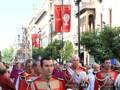 Procesión histórica con uniformes rojos y blancos en una calle decorada con banderas y edificios antiguos.