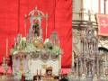 Imagen de una iglesia con un altar adornado con flores y una estatua en el centro, rodeada de velas y un fondo rojo.