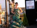 Modela en un desfile de moda flamenca con vestido de flores turquesa y doradas, en un entorno urbano con arquitectura típica de Sevilla.