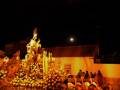 Fiesta religiosa nocturna con una imagen de la Virgen en el centro, rodeada de flores y velas. La luna brilla sobre un edificio blanco con ventanas. Personas observan la imagen, creando una atmósfera festiva y religiosa.