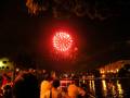 Una explosión de fuegos artificiales rojos ilumina el cielo nocturno, mientras una multitud observa desde la orilla de un río. La celebración parece ser en honor a una festividad especial, con edificios y luces alrededor.
