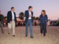 Una persona en un traje azul camina por una playa con otros dos individuos, uno de ellos también en un traje. El entorno es soleado con palmeras y una línea de luces en el fondo, indicando un evento o reunión formal.
