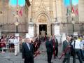 Procesión religiosa frente a la Catedral de Sevilla, con participantes en trajes formales y portando candelabros rojos.