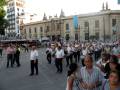 Banda de música desfilando en una plaza histórica con edificios antiguos al fondo.