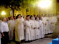 Una procesión religiosa en la noche, con sacerdotes vestidos de blanco y amarillo, posiblemente en una iglesia o plaza pública.
