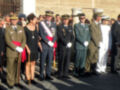 Una imagen de oficiales militares en uniformes formales, posando junto a una mujer en un traje negro. La escena parece ser formal o ceremonial, con el sol brillando sobre ellos y un edificio de fondo.