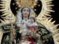 Una venerable escultura de la Virgen María, vestida en trajes tradicionales y sosteniendo un niño Jesús. La escultura está rodeada de velas encendidas y una corona dorada con rayos que simbolizan la divinidad. La escena evoca la devoción y el culto a la Virgen María en contextos religiosos.