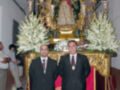 Dos hombres en trajes posan frente a una imagen religiosa dorada, rodeados de flores blancas y velas encendidas.