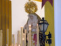 Una estatua religiosa en un interior con velas encendidas y un arco dorado. La estatua parece ser de una figura venerada, posiblemente un santo o virgen, con un manto y tocado. Al fondo hay una bandera roja con letras doradas, probablemente un símbolo religioso o institucional. La atmósfera es reverente y festiva, típica de una celebración religiosa o procesión.
