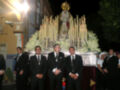 Una procesión religiosa nocturna con una imagen de la Virgen en el centro, rodeada por velas encendidas y flores. Un grupo de hombres en trajes formales, algunos con insignias militares, observa la imagen. La escena se desarrolla en un entorno urbano con edificios y árboles al fondo.